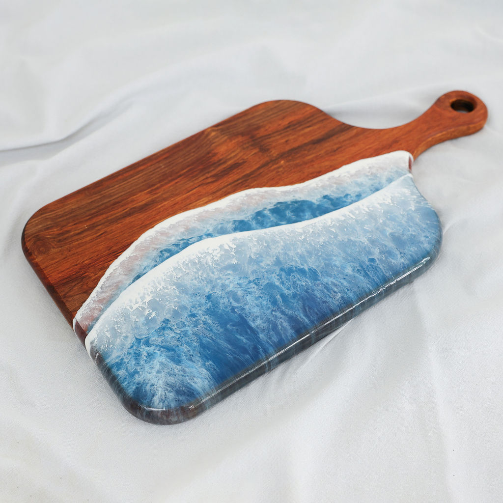 Angled view of handcrafted kiaat wood medium paddle serving board with blue ocean resin wave design